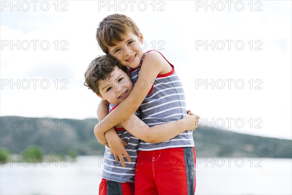 Brothers (4-5, 6-7) embracing on beach by lake