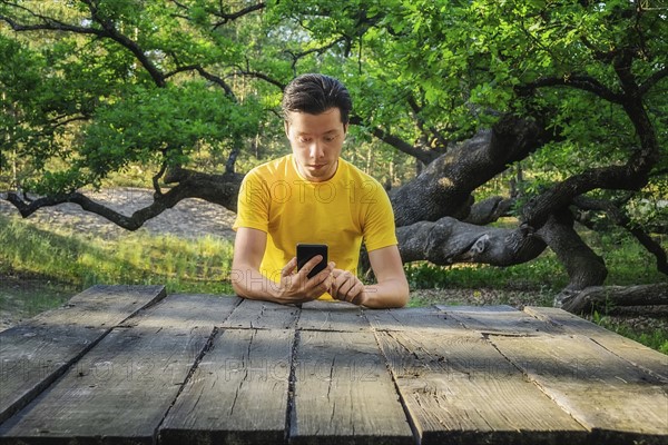 Ukraine, Dnepropetrovsk region, Novomoskovsk district, Man sitting at wooden table and using smart phone with Oak (Quercus) tree in background