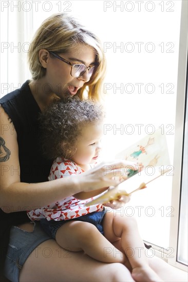 Mother sitting with daughter (12-17 months) on windowsill and reading book