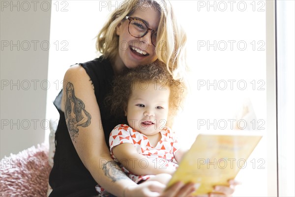 Mother sitting with daughter (12-17 months) on windowsill and reading book