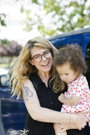 Mother carrying daughter (12-17 months) in front of car