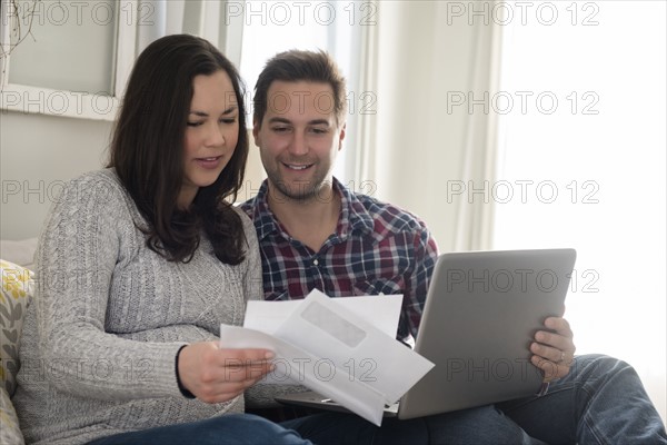 Mid adult couple sitting on sofa and looking at letter, man holding laptop