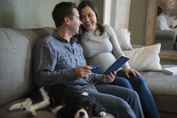 Mid adult couple shopping online in living room, sitting on sofa with puppy
