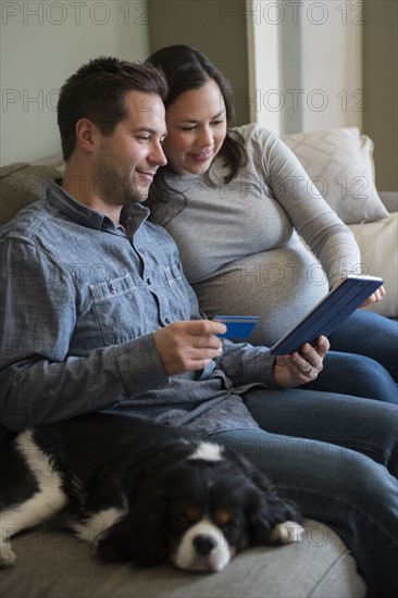 Mid adult couple shopping online in living room, sitting on sofa with puppy