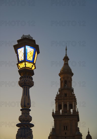 Spain, Andalusia, Seville, Plaza de Espana, Illuminated old fashioned street lamp and tower at dawn