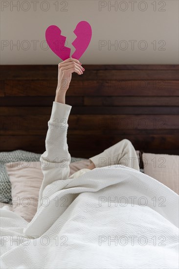 Woman holding aloft pink broken heart while lying in bed