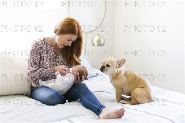 Dog sitting by woman holding her newborn son on bed