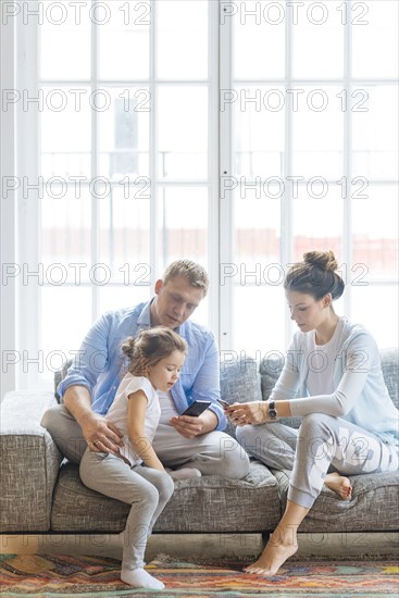 Parents holding smart phones on sofa with their daughter