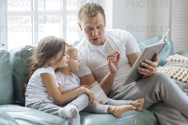 Father reading book to daughters on sofa