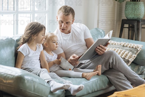 Father reading book to daughters on sofa