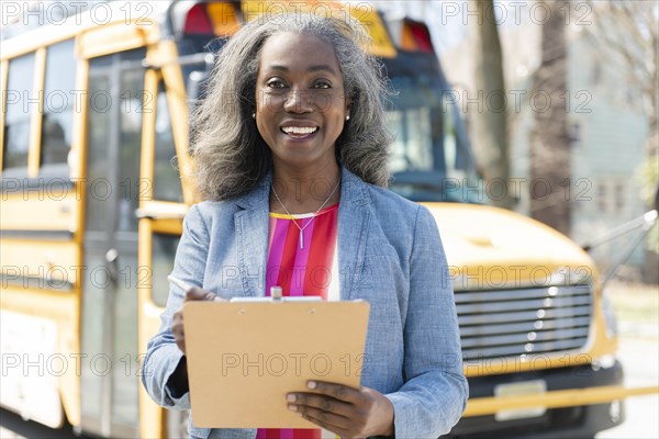 Smiling teacher with clipboard by school bus