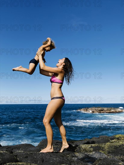 Mother lifting her son on rocks by sea