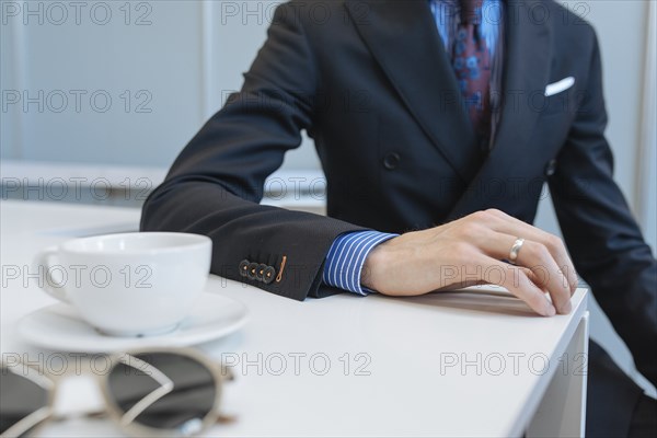 Man wearing suit leaning on table by tea cup and sunglasses