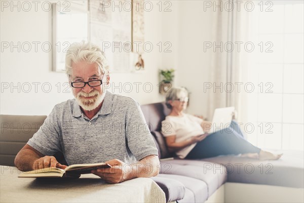 Senior man reading book as his wife uses laptop in living room