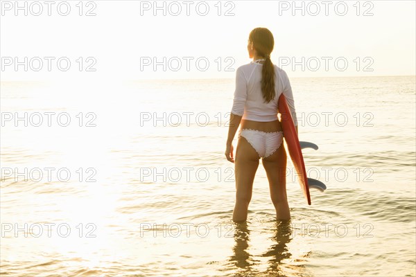 Woman holding surfboard in sea at sunset