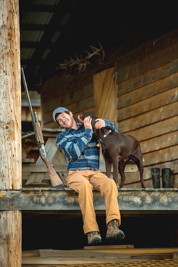 Man petting chocolate Labrador on porch of log cabin