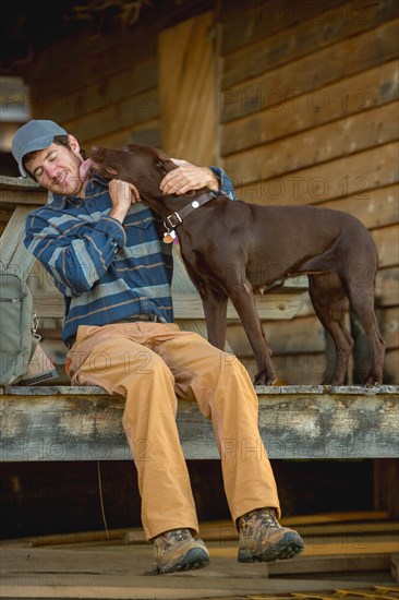 Chocolate Labrador licking man's face on porch of log cabin