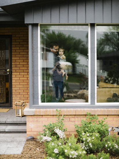 Father holding his daughter behind window of house