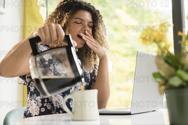 Tired woman pouring coffee by laptop