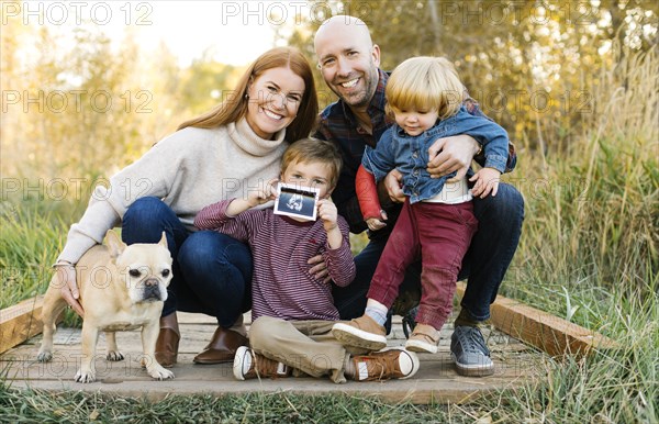 Smiling family with ultrasound photograph and pet dog on forest boardwalk