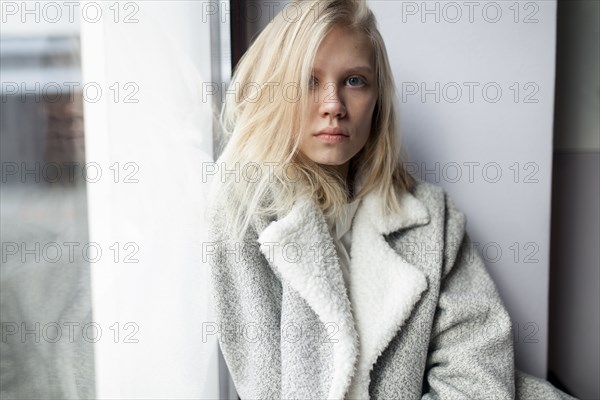 young woman in wool coat leaning against wall by window