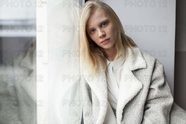 young woman in wool coat leaning against wall by window