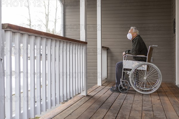 Senior man in wheelchair wearing protective mask to prevent coronavirus transmission on porch