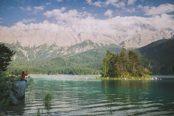 Germany, Bavaria, Eibsee, Young woman sitting on rock by Eibsee Lake in Bavarian Alps and photographing landscape with smart phone