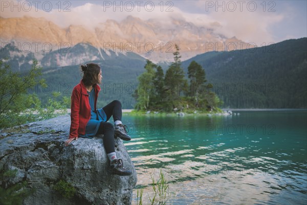 Germany, Bavaria, Eibsee, Young woman sitting on rock and looking at scenic view by Eibsee lake in Bavarian Alps