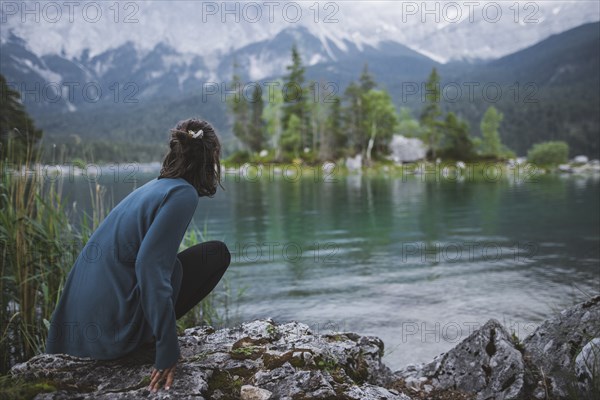 Germany, Bavaria, Eibsee, Young woman crouching on rock at shore of Eibsee lake in Bavarian Alps