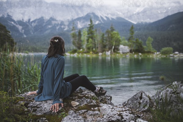 Germany, Bavaria, Eibsee, Young woman sitting on rock at shore of Eibsee lake in Bavarian Alps