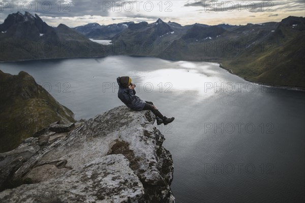 Norway, Senja, Man taking photo sitting on edge of steep cliff on top of mountain Segla