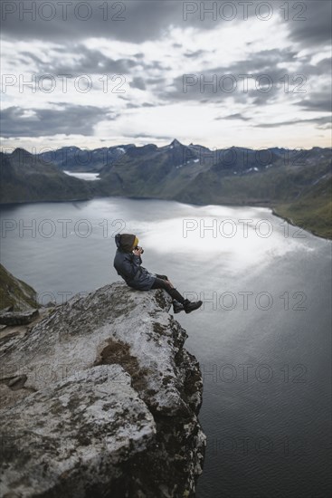 Norway, Senja, Man taking photo sitting on edge of steep cliff on top of mountain Segla
