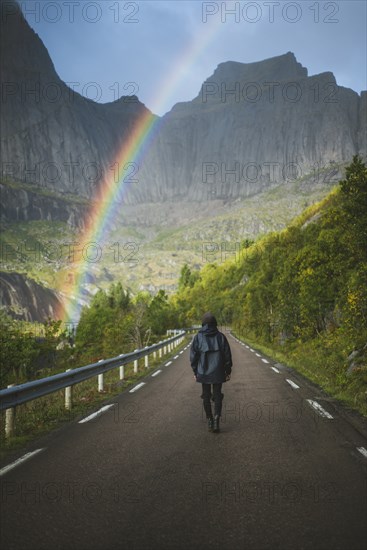 Norway, Lofoten Islands, Man walking down road with mountains and rainbow in background