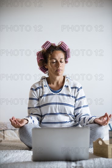 Woman with hair curlers meditating on bed in front of laptop