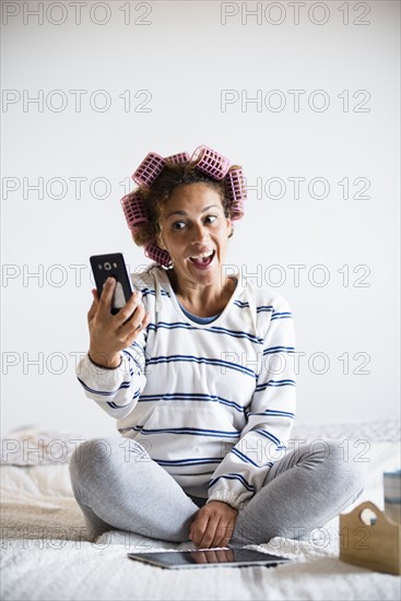 Woman with hair curlers sitting on bed and taking selfie