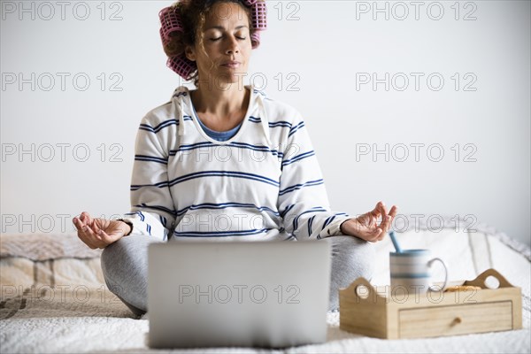 Woman with hair curlers meditating on bed in front of laptop