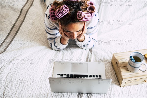 Woman with hair curlers lying on bed and looking at laptop