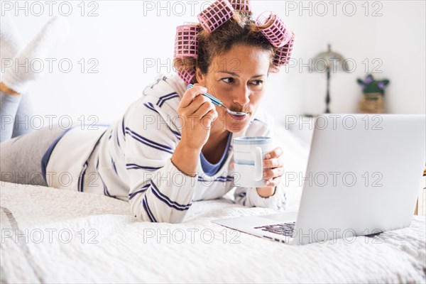 Woman with hair curlers lying on bed and looking at laptop