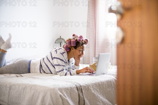 Woman with hair curlers lying on bed and using laptop