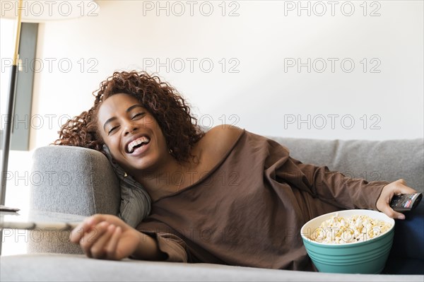 Laughing woman on sofa with TV remote and popcorn