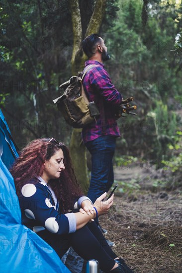 Woman sitting in tent using smart phone by man holding sticks