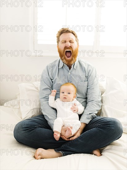 Yawning father sitting on bed with baby boy (2-3 months)