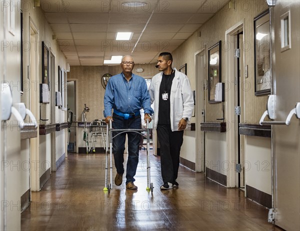 Male nurse assisting senior man walking down corridor with mobility walker