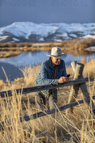 USA, Idaho, Sun Valley, Senior man in cowboy hat leaning against fence texting