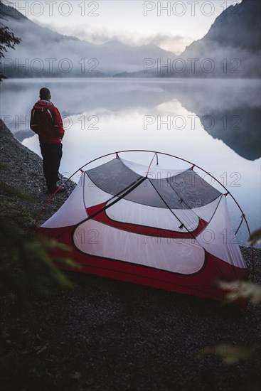 Austria, Plansee, Young man standing near tent and looking at Plansee lake at sunrise
