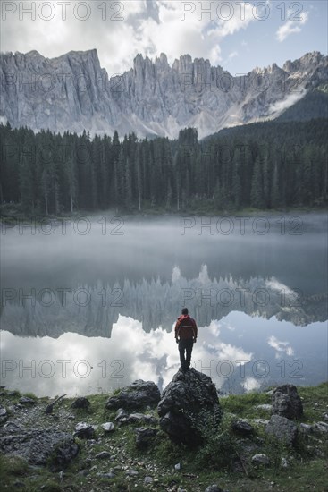 Italy, Carezza, Young man standing on rock at Lago di Carezza in Dolomite Alps at dawn