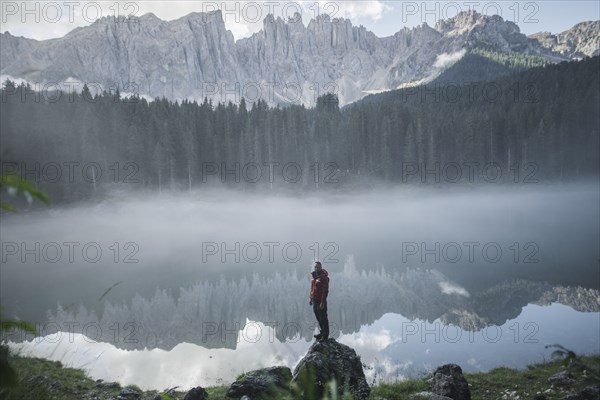 Italy, Carezza, Young man standing on rock at Lago di Carezza in Dolomite Alps at dawn