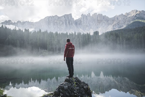 Italy, Carezza, Young man standing on rock and looking at Lago di Carezza in Dolomite Alps at dawn