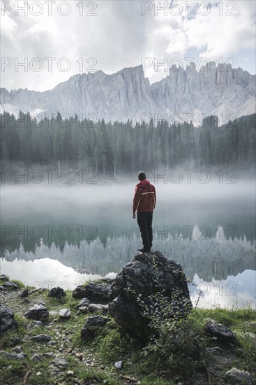 Italy, Carezza, Young man standing on rock at Lago di Carezza in Dolomite Alps at dawn